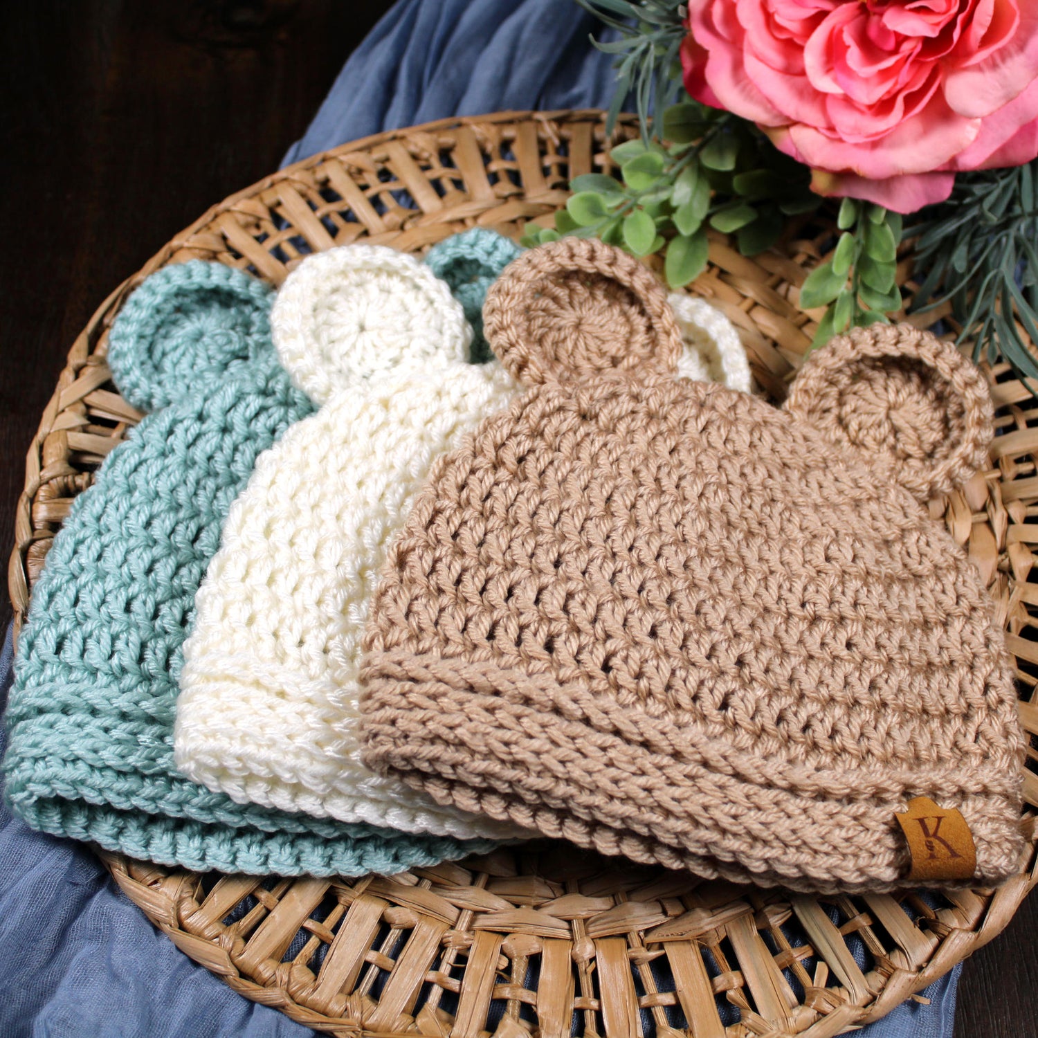 tack of handmade crochet baby hats in green, cream, and taupe topped with a pink flower.