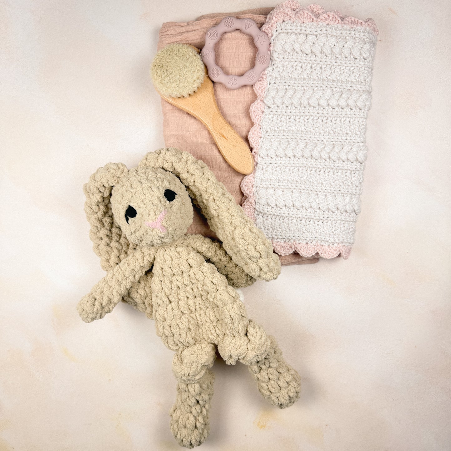 Flat lay of the Petal & Pearl Keepsake Edit featuring a plush bunny, crochet security blanket, muslin swaddle, wooden baby brush, and silicone teether.