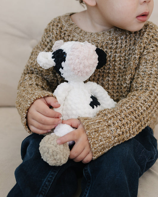 A young child in a textured tan sweater holding a white and black handcrafted cow plush in a neutral, modern home setting.