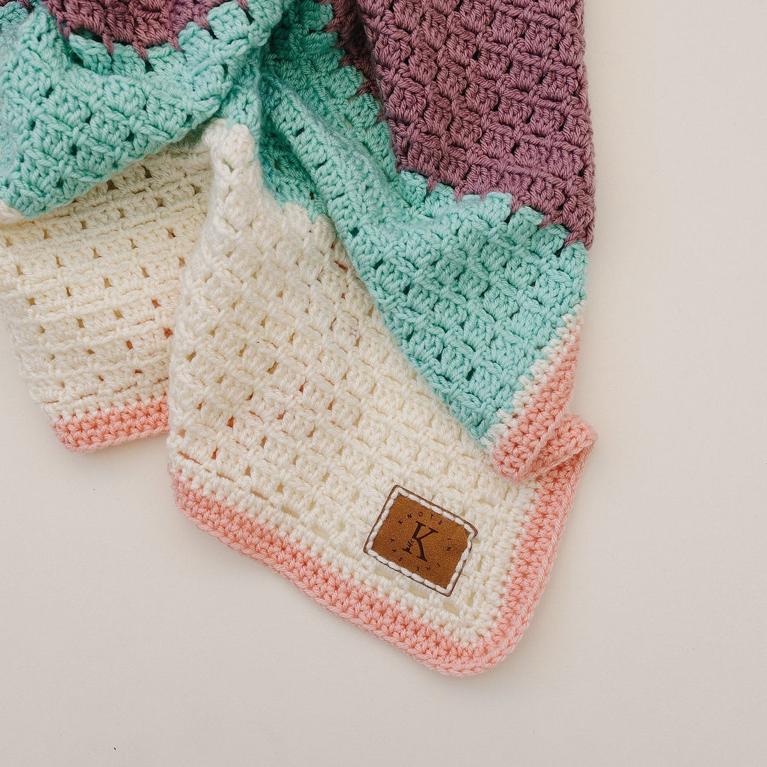 Close-up of a handmade crochet blanket with cream, sage, and blush stripes featuring a leather "K" brand tag.