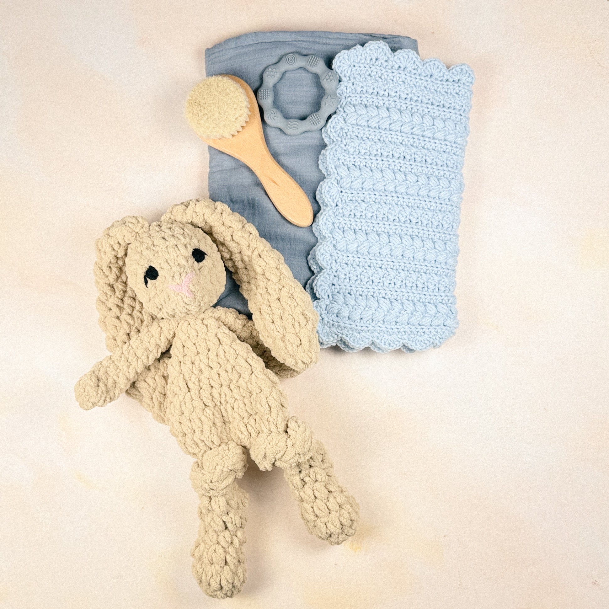 Flat lay of the Stone Blue Keepsake Edit featuring a plush bunny, crochet security blanket, muslin swaddle, wooden baby brush, and silicone teether.