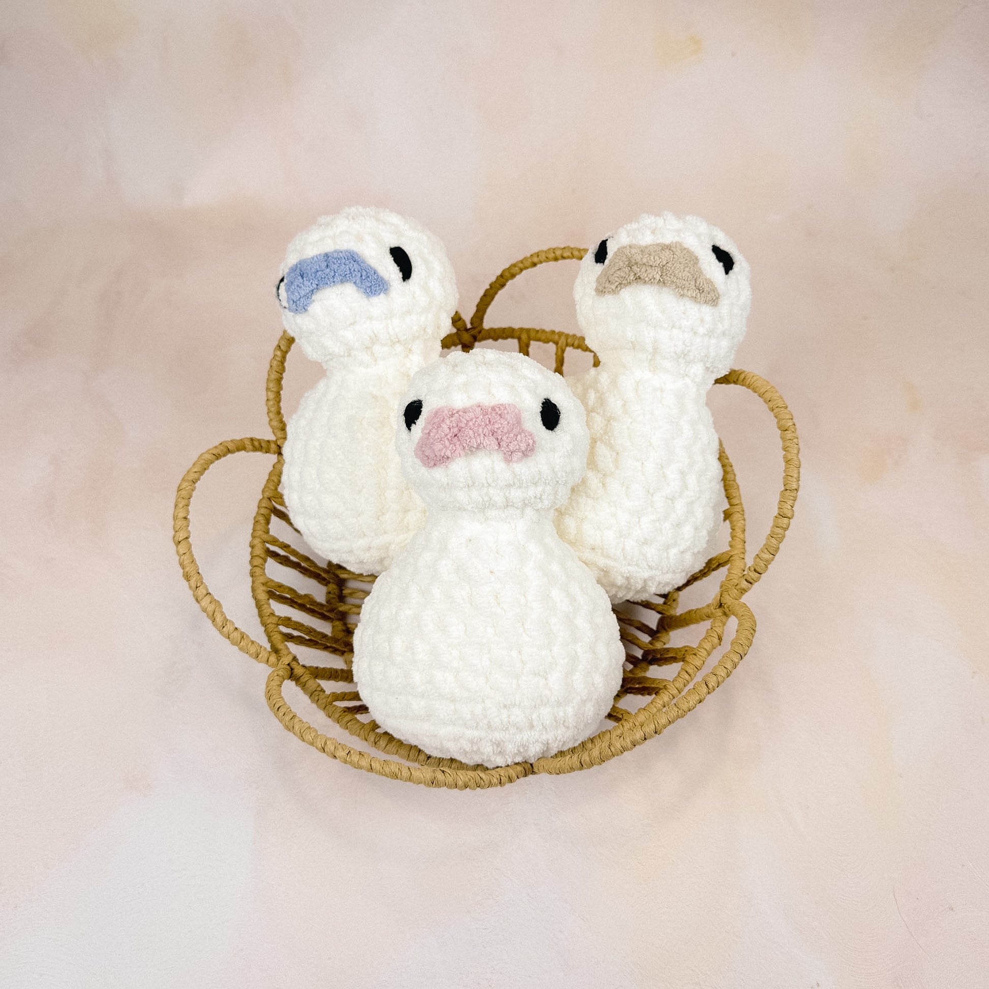 Three crochet duck plush toys with blush, taupe, and blue beaks displayed in a woven basket against a neutral backdrop.